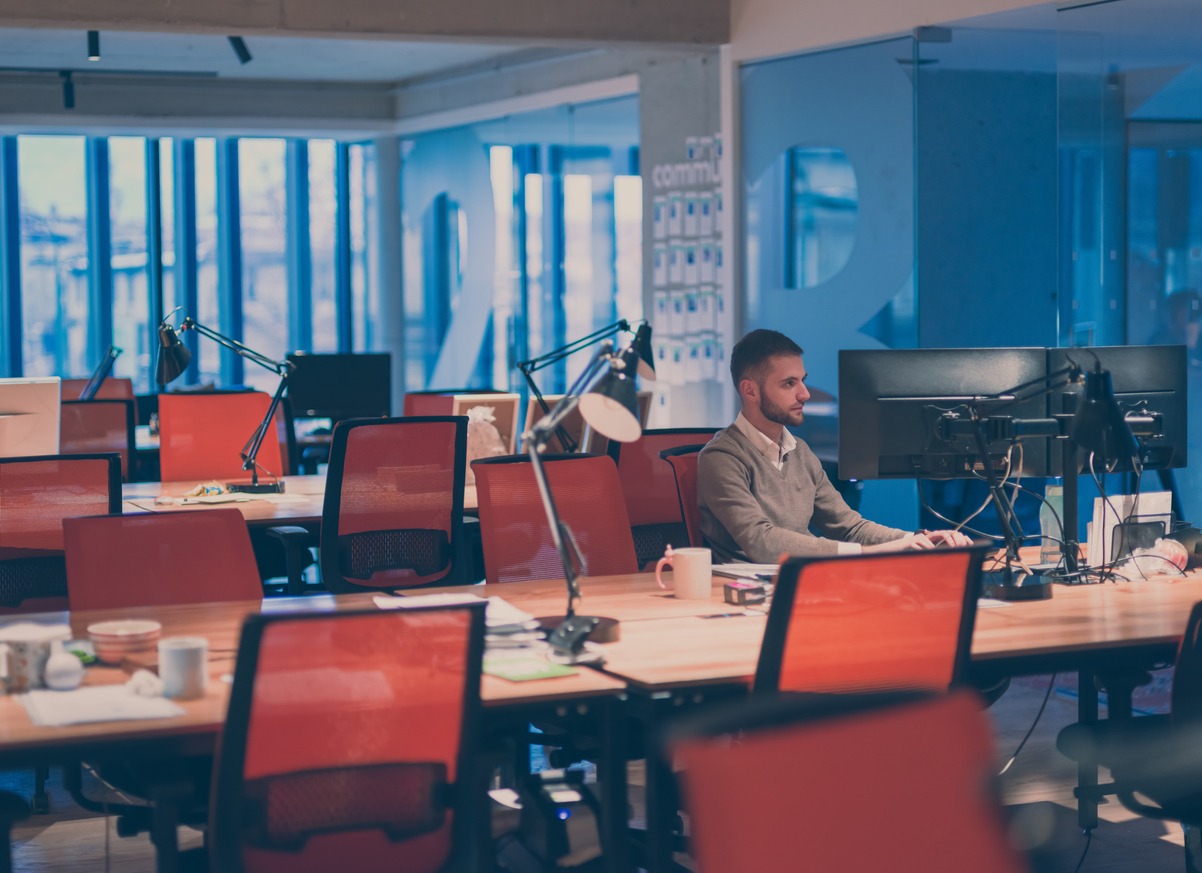 Young businessman working overtime alone at his desk on desktop computer in an open space modern coworking office late at night with city lights glowing in the background. High quality photo